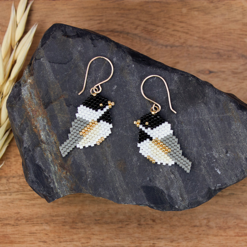 A pair of handmade beaded bird drop earrings with gold-tone fishhook wires, displayed on a dark slate rock over a wooden surface.