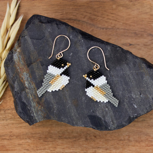 A pair of handmade beaded bird drop earrings with gold-tone fishhook wires, displayed on a dark slate rock over a wooden surface.