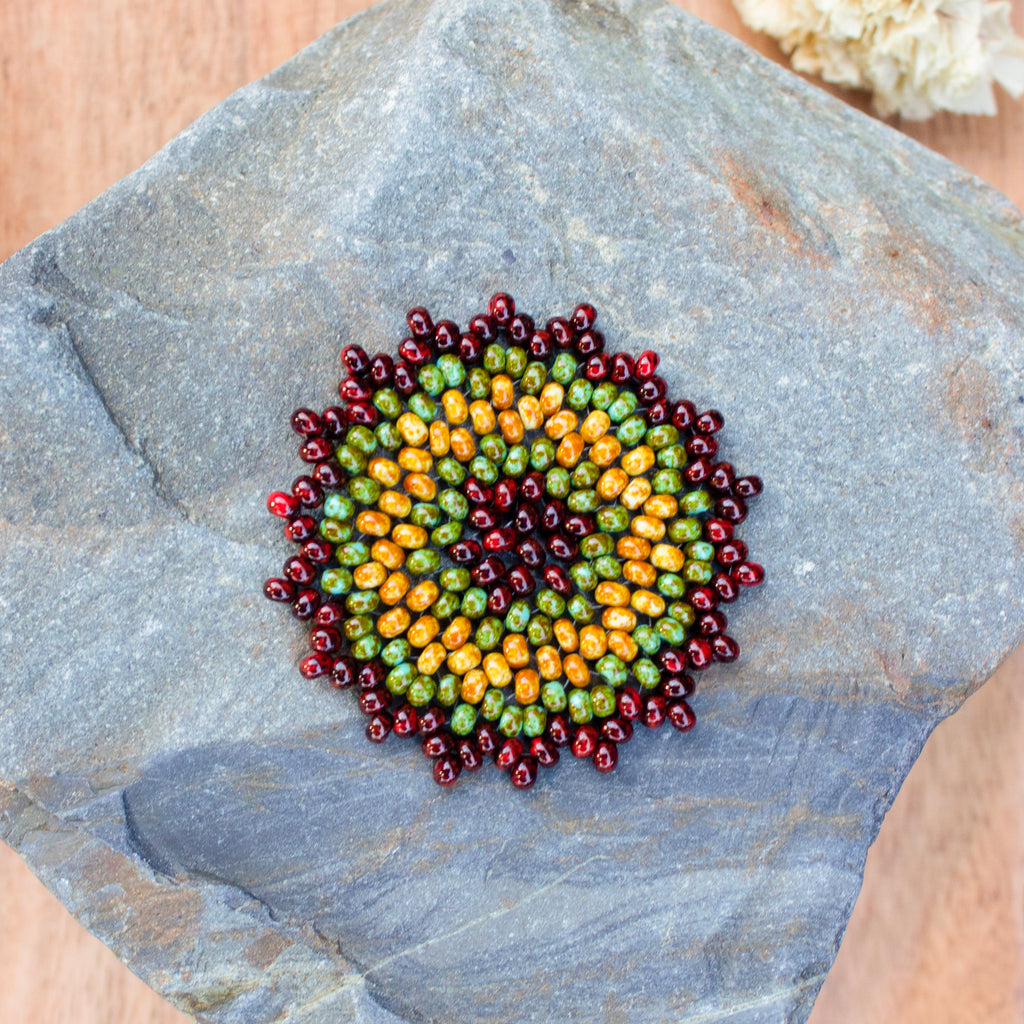 A circular beaded medallion with concentric rings of red, green, and orange beads, displayed on a gray slate stone. A sensory tool