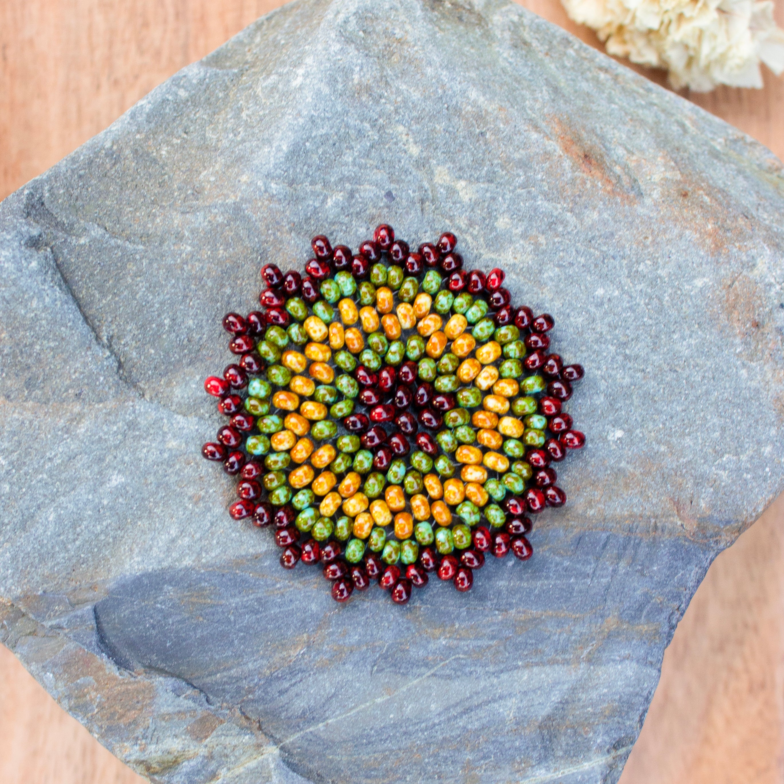 A circular beaded medallion with concentric rings of red, green, and orange beads, displayed on a gray slate stone. A sensory tool