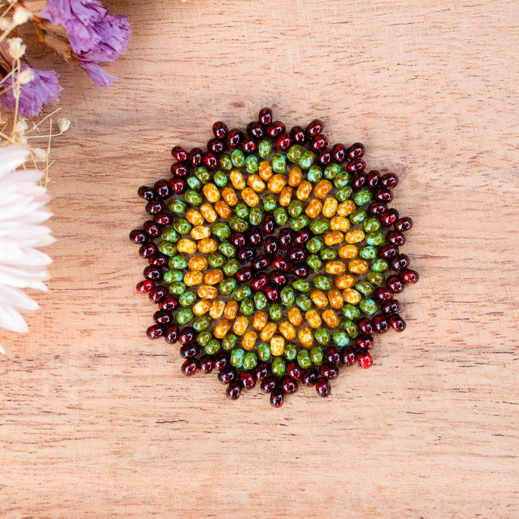 A circular arrangement of small seed beads forming a sunburst pattern, placed on a light wood surface with dried flowers in the top-left corner. a Sensory tool