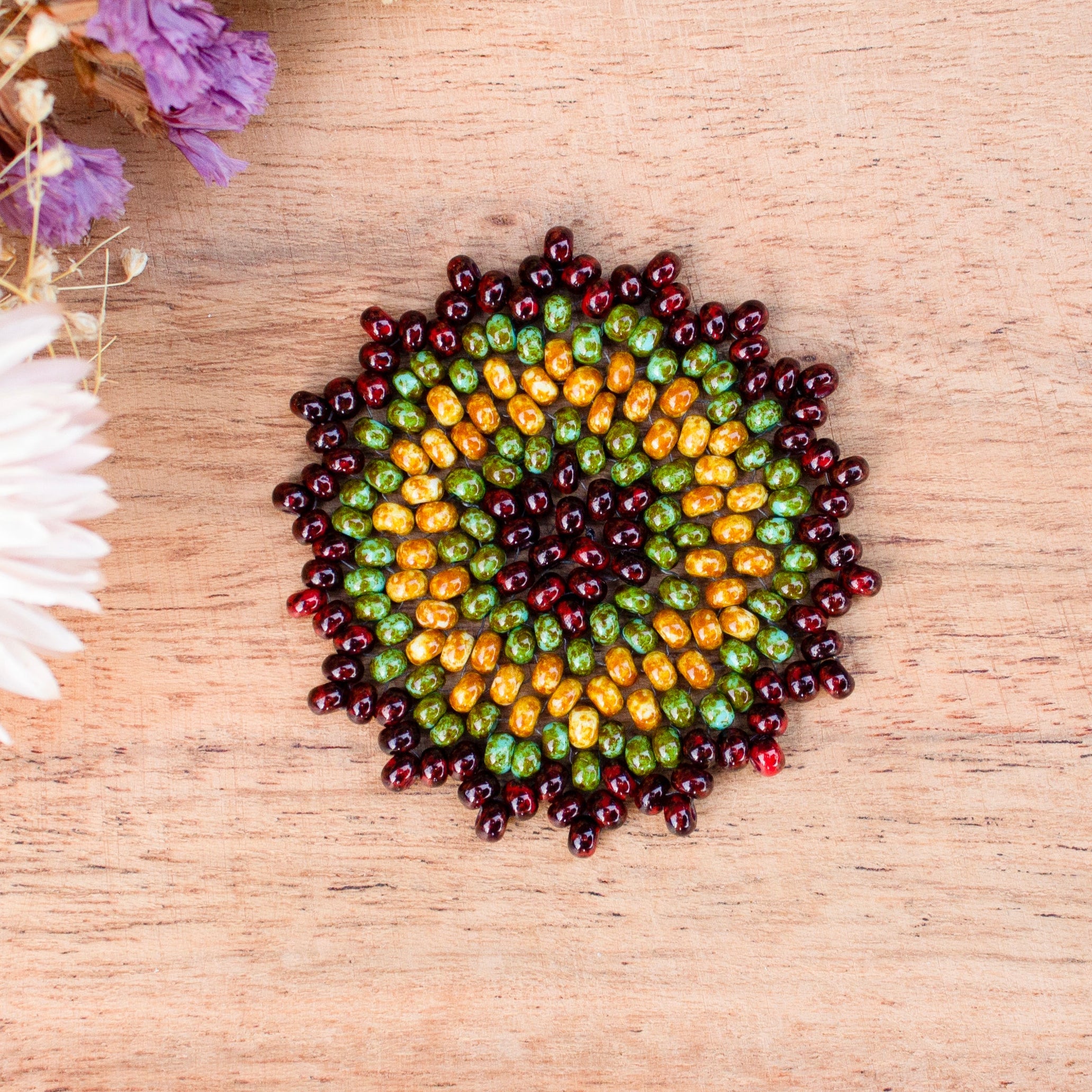 A circular arrangement of small seed beads forming a sunburst pattern, placed on a light wood surface with dried flowers in the top-left corner. a Sensory tool