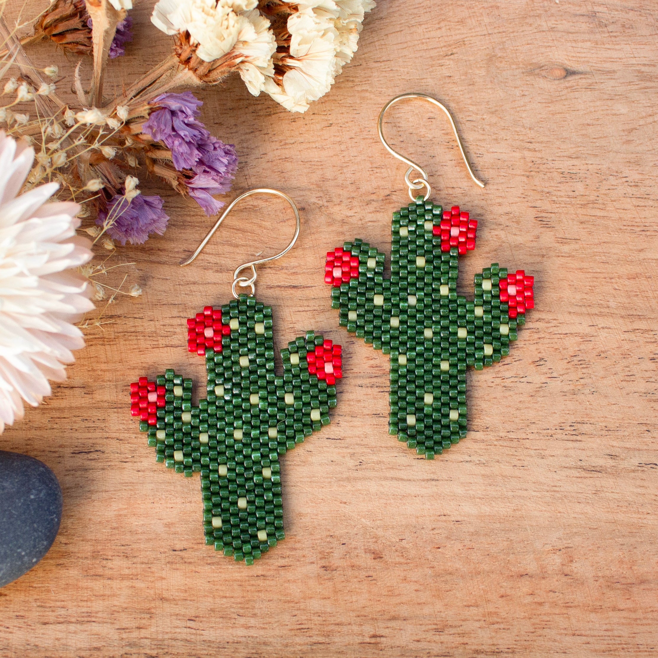A pair of handmade beaded cactus earrings with green seed beads, red flower accents, and gold fishhook wires, displayed on a wooden surface next to dried flowers.