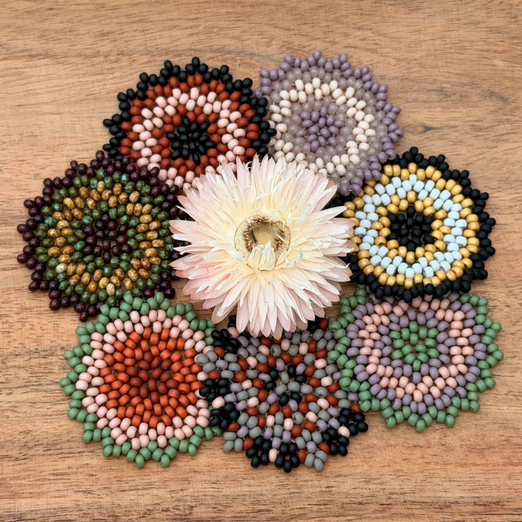 Seven circular flower-shaped bead arrangements made from seed beads, arranged in a circle around a pale pink flower on a wooden surface. The beads come in many colors including green, blue, yellow, purple, black, white, orange, and brown.