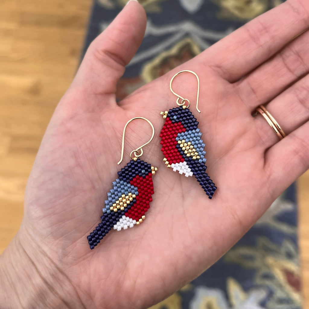 A pair of handmade beaded bird drop earrings held in a person's hand. The earrings are designed to look like birds, featuring red, navy blue, light blue, white, and gold seed beads, with gold fishhook wires for the ears.