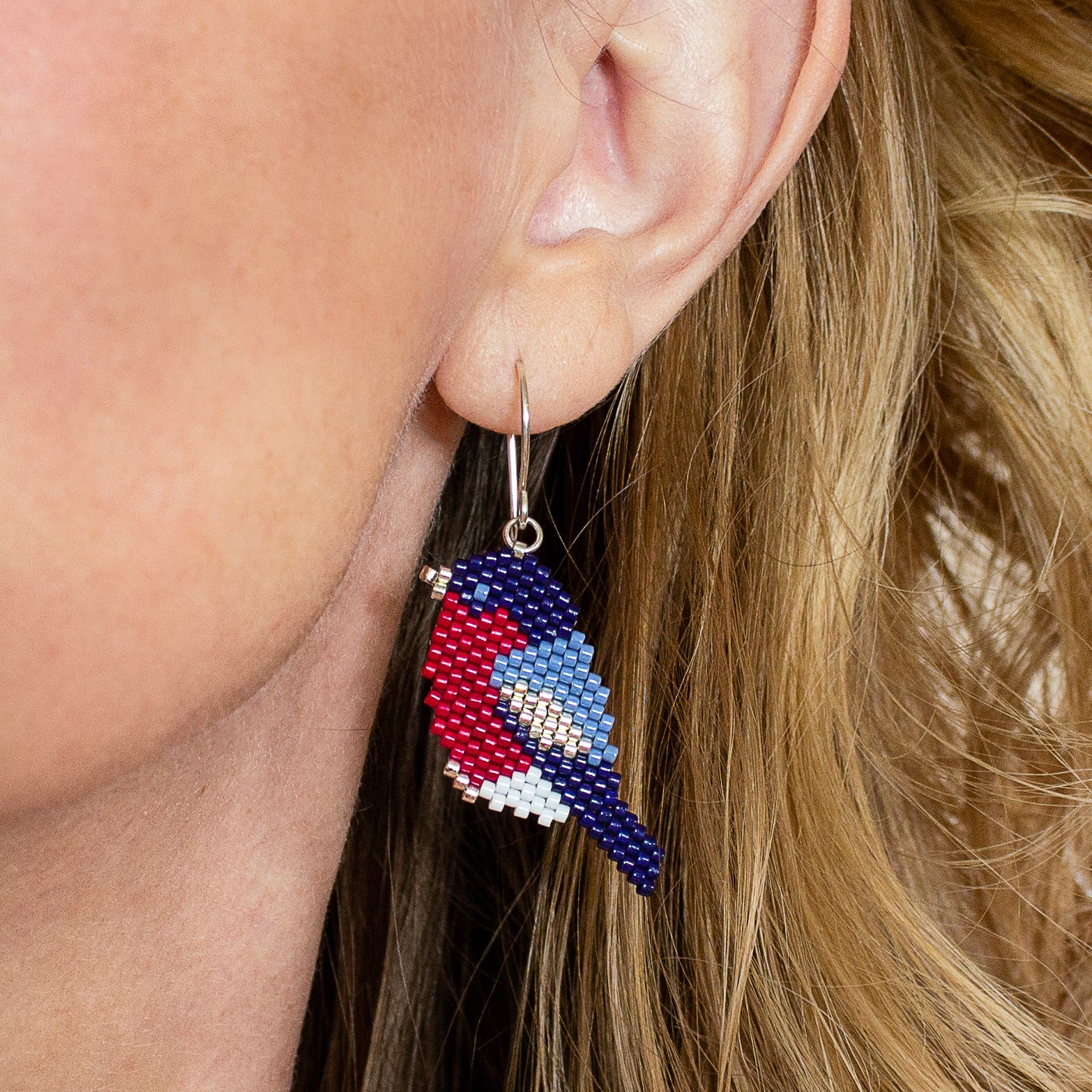 A close-up of a person's ear wearing a beaded bird drop earring. The earring features a bird design in red, blue, white, and light blue, suspended from a silver fishhook ear wire.