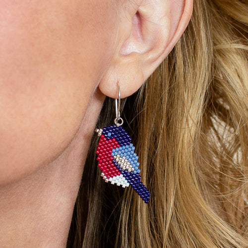 A close-up of a person's ear wearing a beaded bird drop earring. The earring features a bird design in red, blue, white, and light blue, suspended from a silver fishhook ear wire.