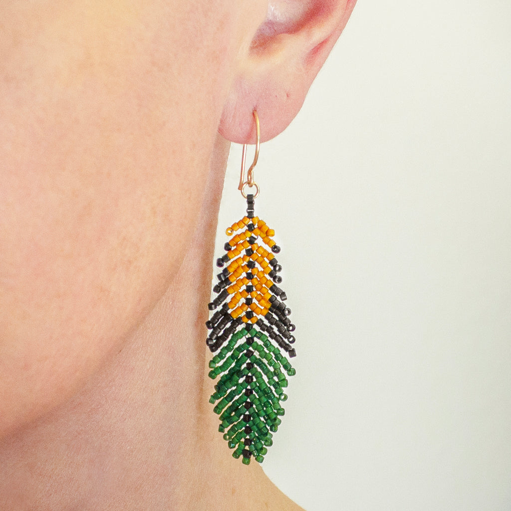 A close-up of a person's ear wearing a long beaded feather drop earring in green, yellow, and black with a gold-tone hook.