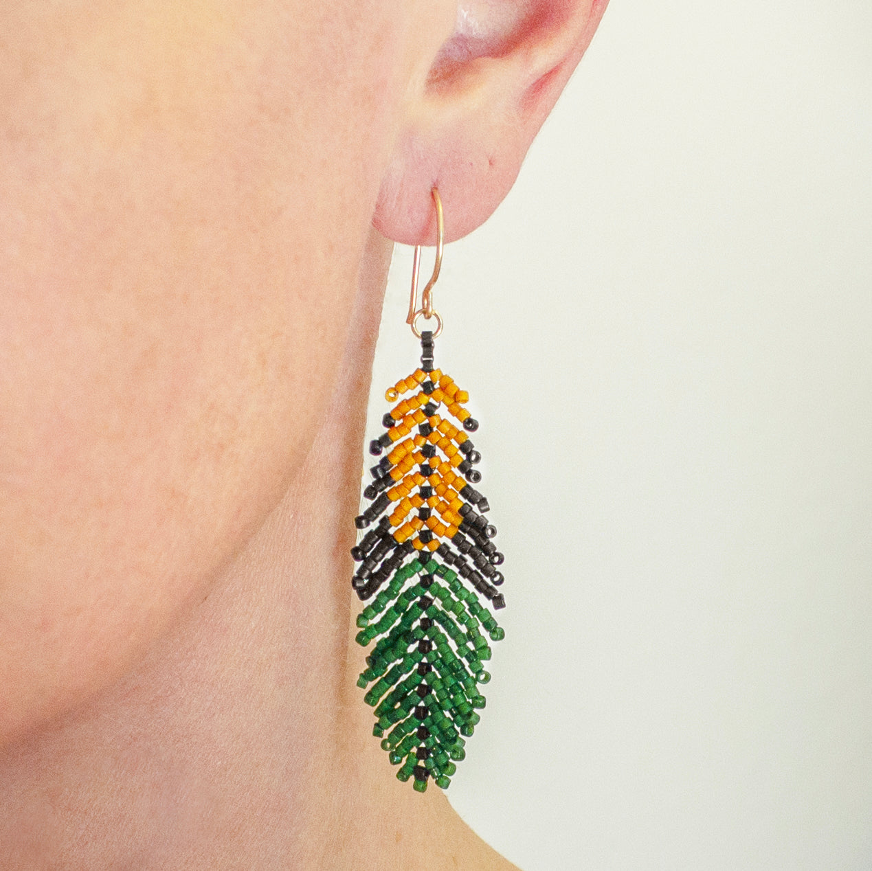 A close-up of a person's ear wearing a long beaded feather drop earring in green, yellow, and black with a gold-tone hook.