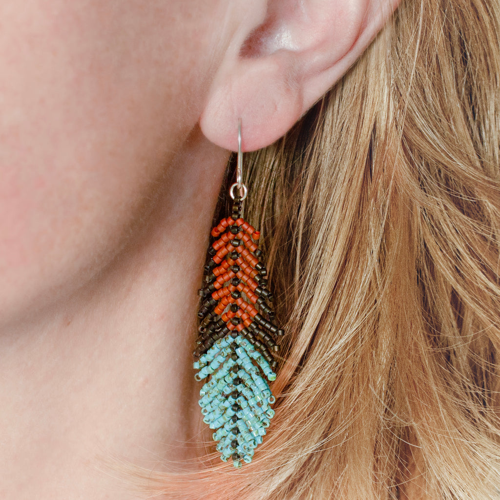 A close-up of a person's ear wearing a dangling beaded feather earring. The earring features a dark brown top section with orange accents, and a light blue feather-shaped bottom section, hanging against blonde hair.
