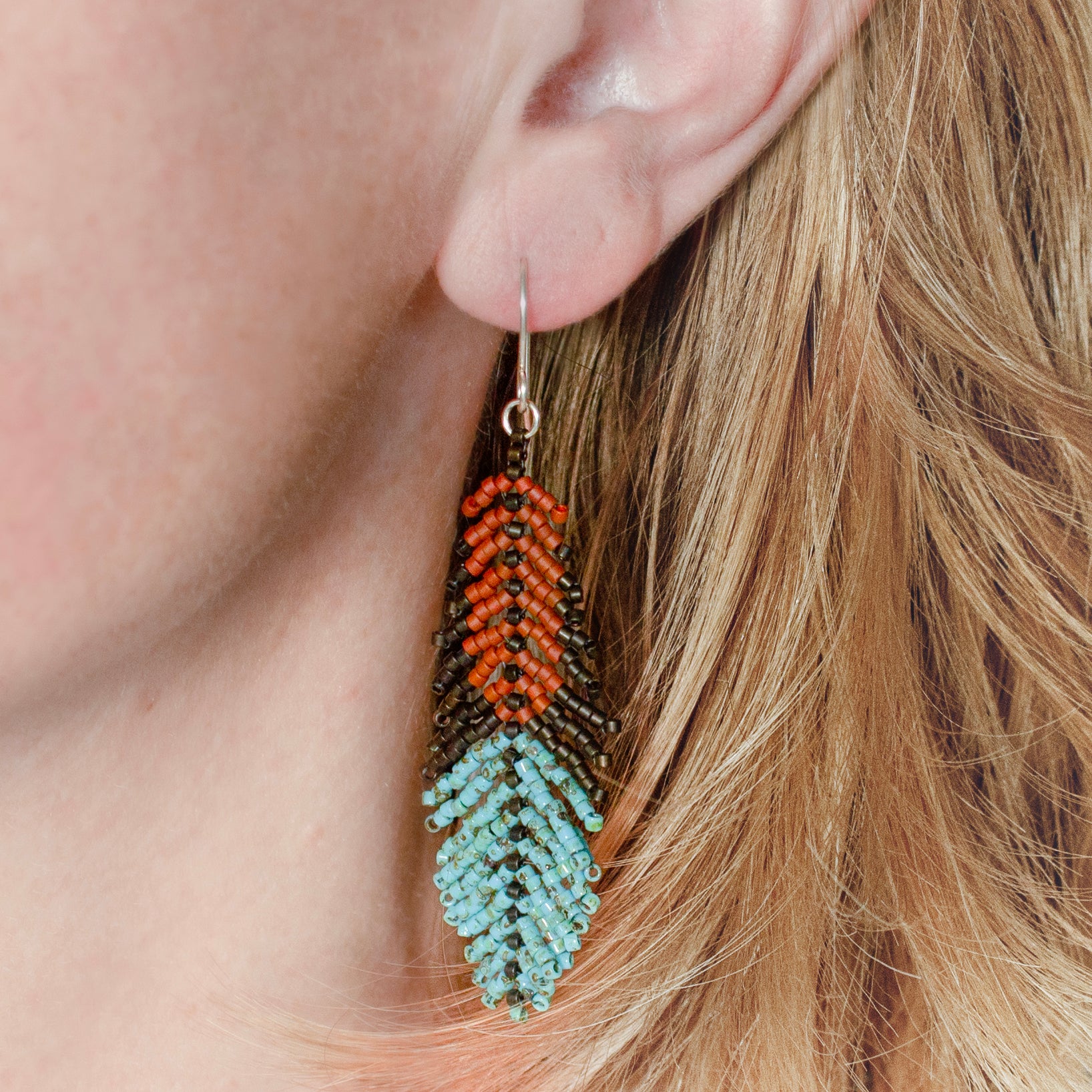 A close-up of a person's ear wearing a dangling beaded feather earring. The earring features a dark brown top section with orange accents, and a light blue feather-shaped bottom section, hanging against blonde hair.