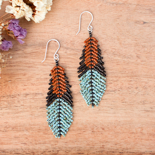 A pair of handmade beaded feather drop earrings with silver fishhook wires, displayed on a light brown wooden surface with dried flowers in the corner.