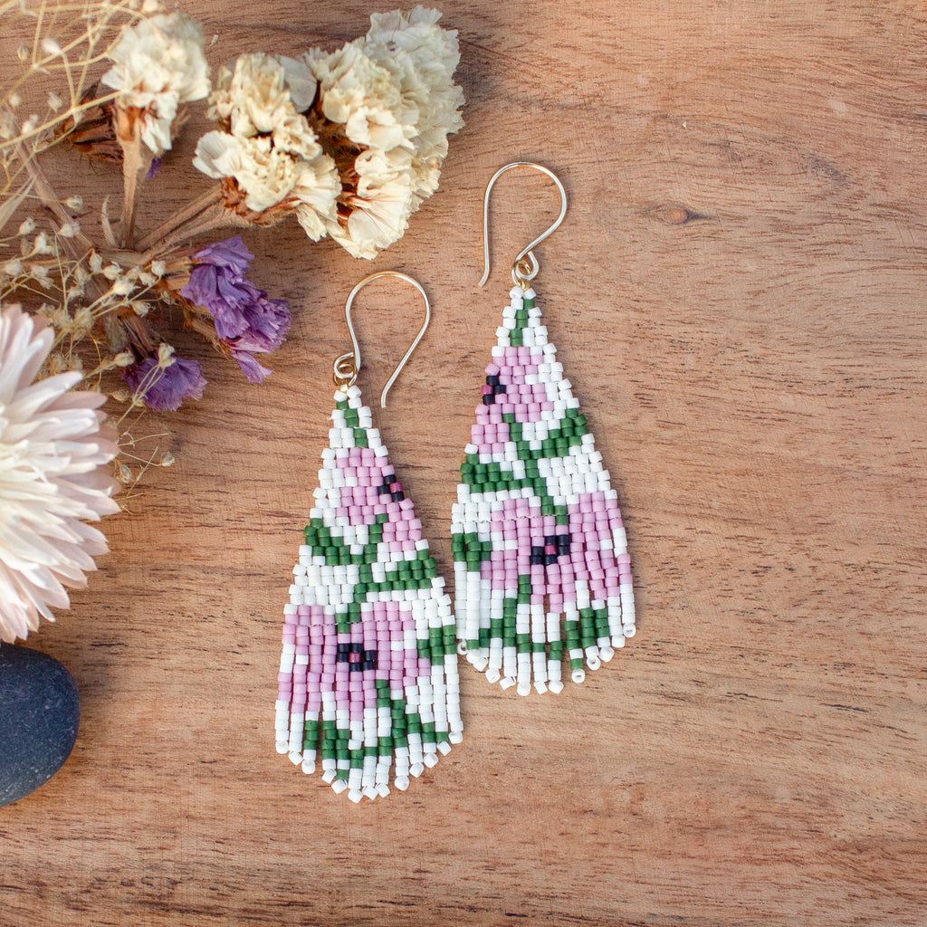 A pair of teardrop-shaped beaded dangle earrings with a floral pattern in pink, green, and white, hanging from gold-tone fishhook wires on a wooden surface with dried flowers as props.