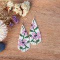 A pair of teardrop-shaped beaded dangle earrings with a floral pattern in pink, green, and white, hanging from gold-tone fishhook wires on a wooden surface with dried flowers as props.