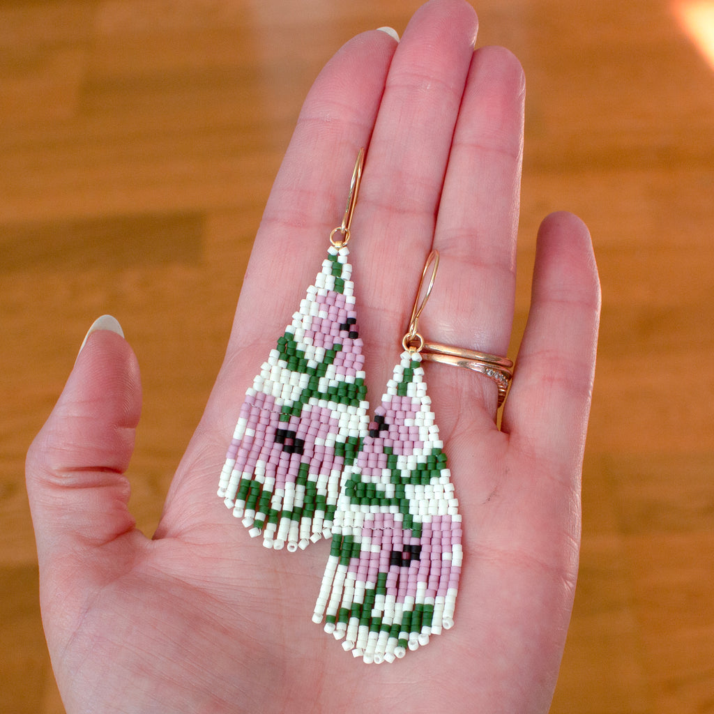 A pair of handmade beaded teardrop earrings with a pink and green floral pattern, held in a person's hand against a wooden background.