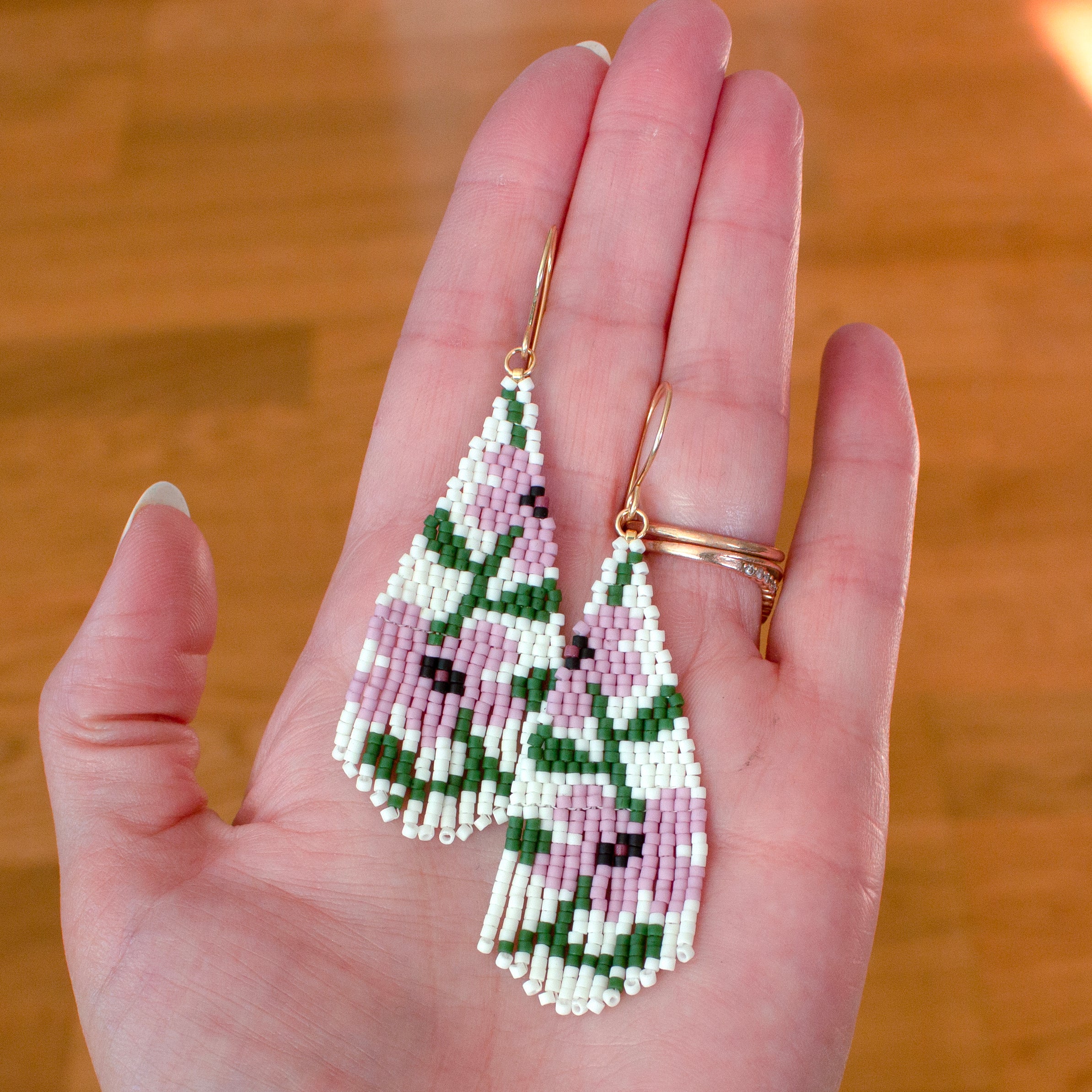 A pair of handmade beaded teardrop earrings with a pink and green floral pattern, held in a person's hand against a wooden background.