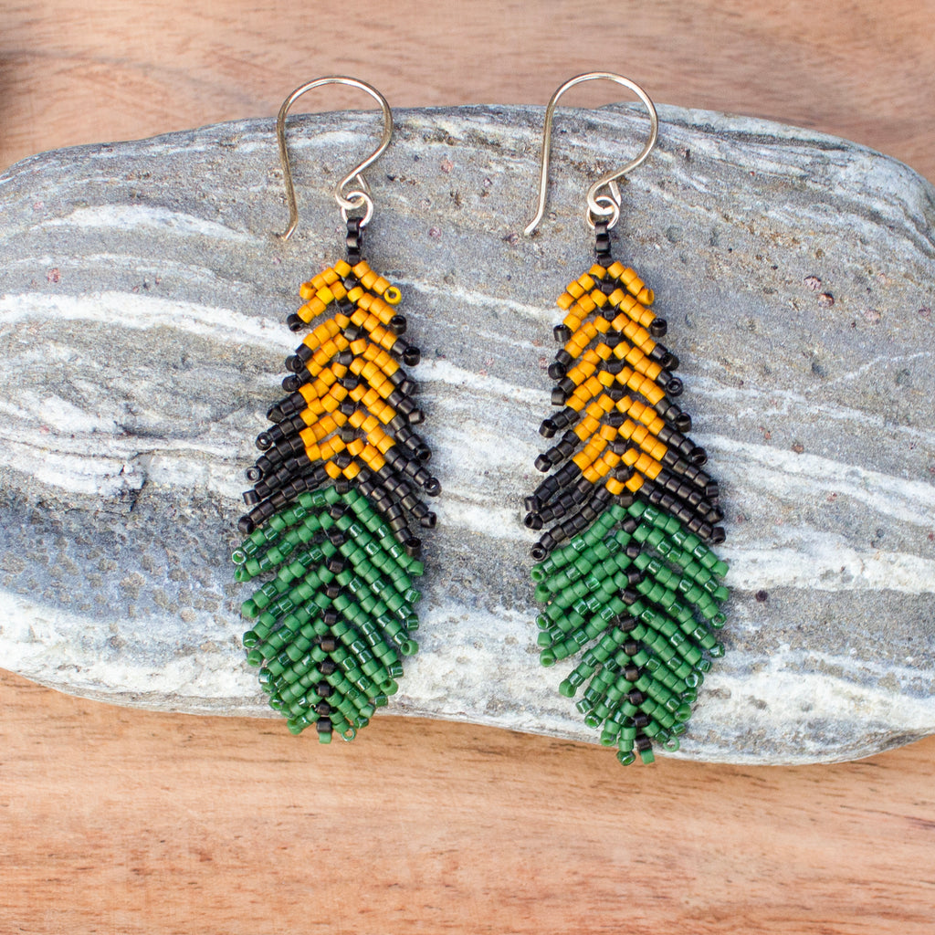 A pair of handmade beaded dangle earrings shaped like feathers, featuring yellow and black upper sections and green lower sections, displayed on a gray stone over a wooden surface.