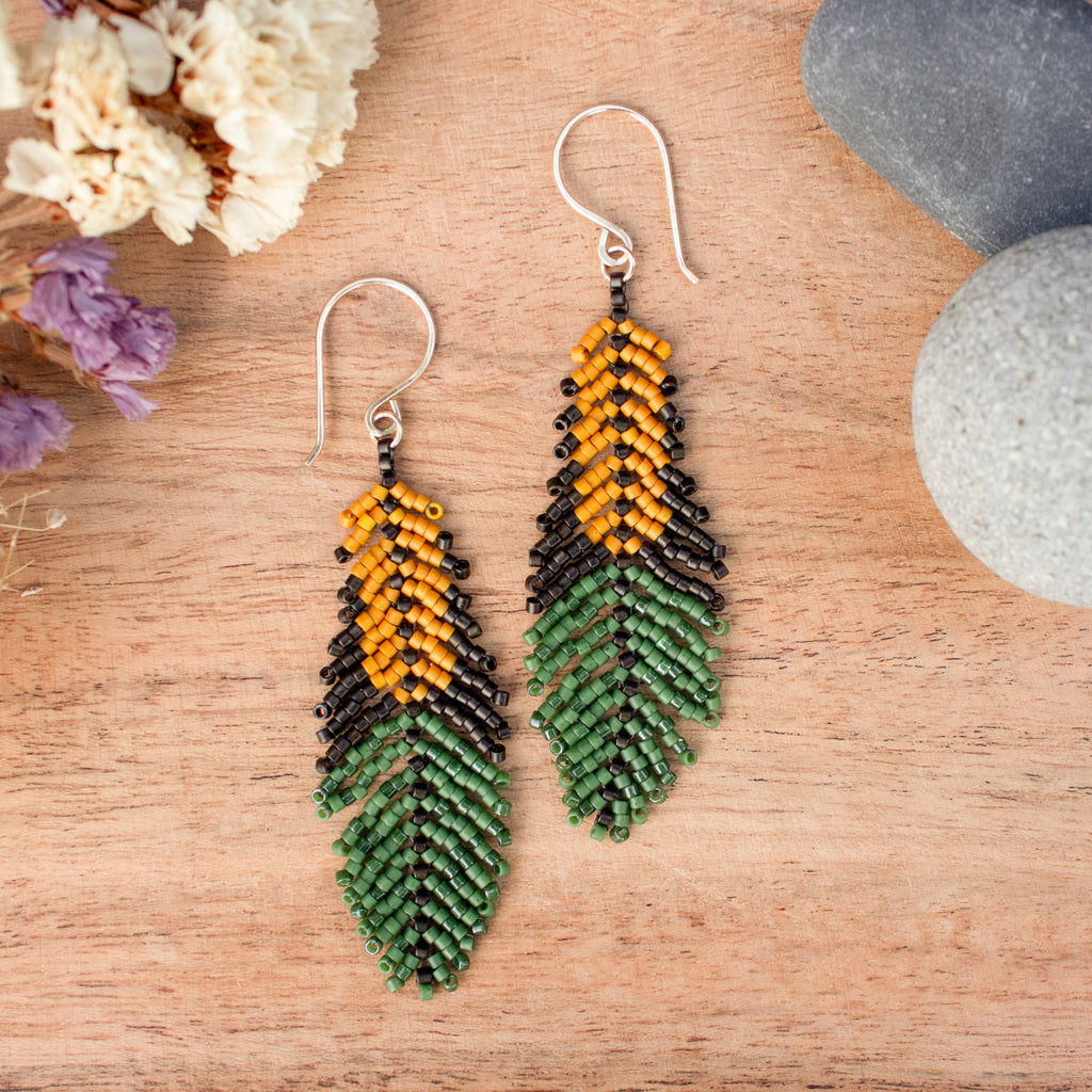 A pair of handmade beaded drop earrings shaped like feathers, featuring a yellow top section, a black middle section, and a green bottom section, hanging on silver fishhook wires over a wooden surface with dried flowers and stones.