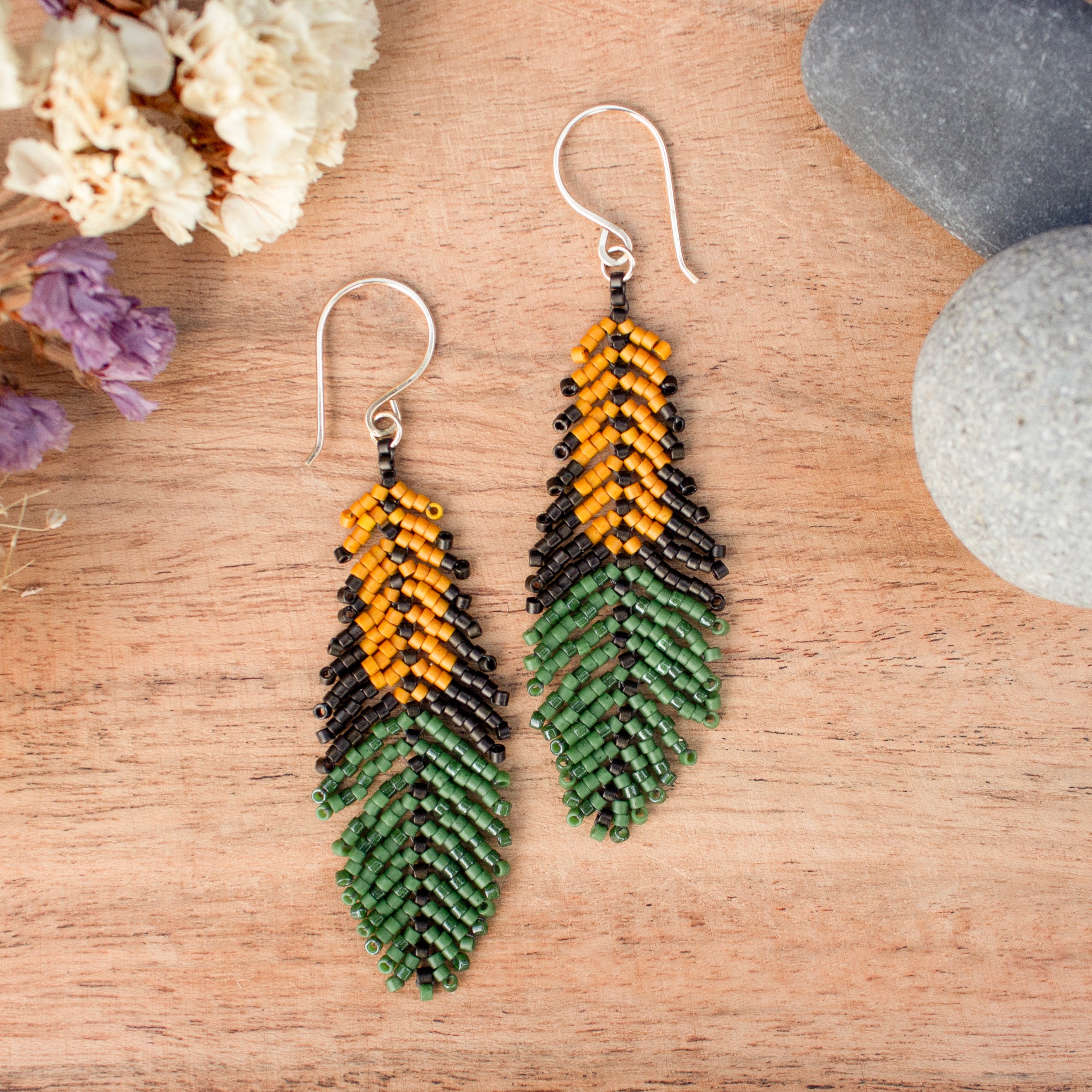 A pair of handmade beaded drop earrings shaped like feathers, featuring a yellow top section, a black middle section, and a green bottom section, hanging on silver fishhook wires over a wooden surface with dried flowers and stones.