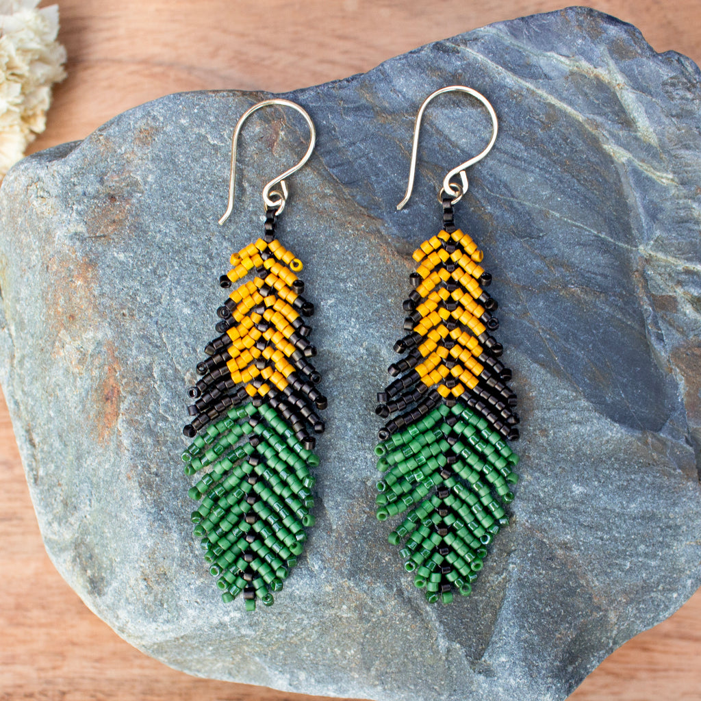 A pair of handmade beaded earrings shaped like corn kernels and leaves, displayed on a gray stone. The earrings feature yellow and black seed beads at the top and green seed beads at the bottom, suspended from silver fishhook wires.