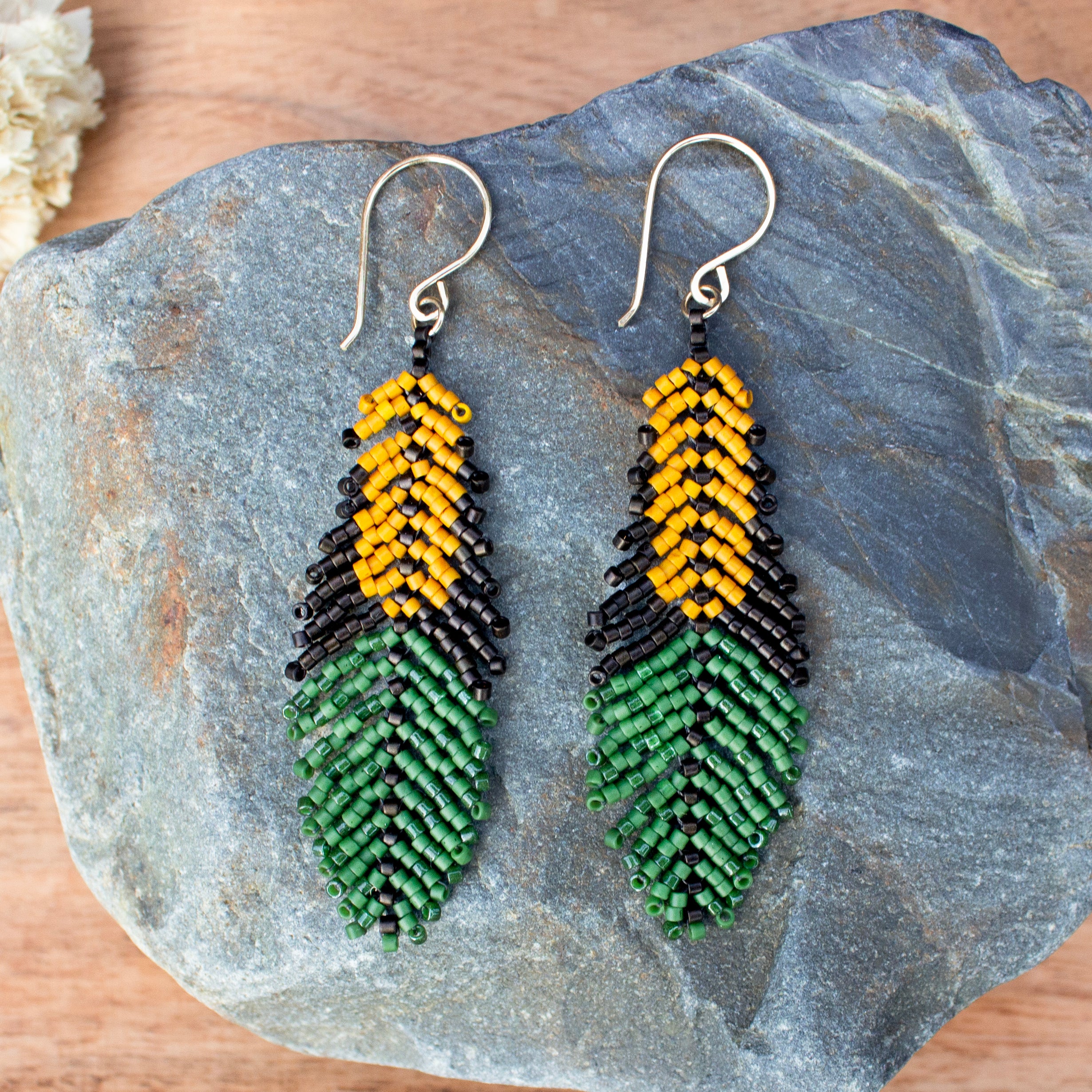A pair of handmade beaded earrings shaped like corn kernels and leaves, displayed on a gray stone. The earrings feature yellow and black seed beads at the top and green seed beads at the bottom, suspended from silver fishhook wires.