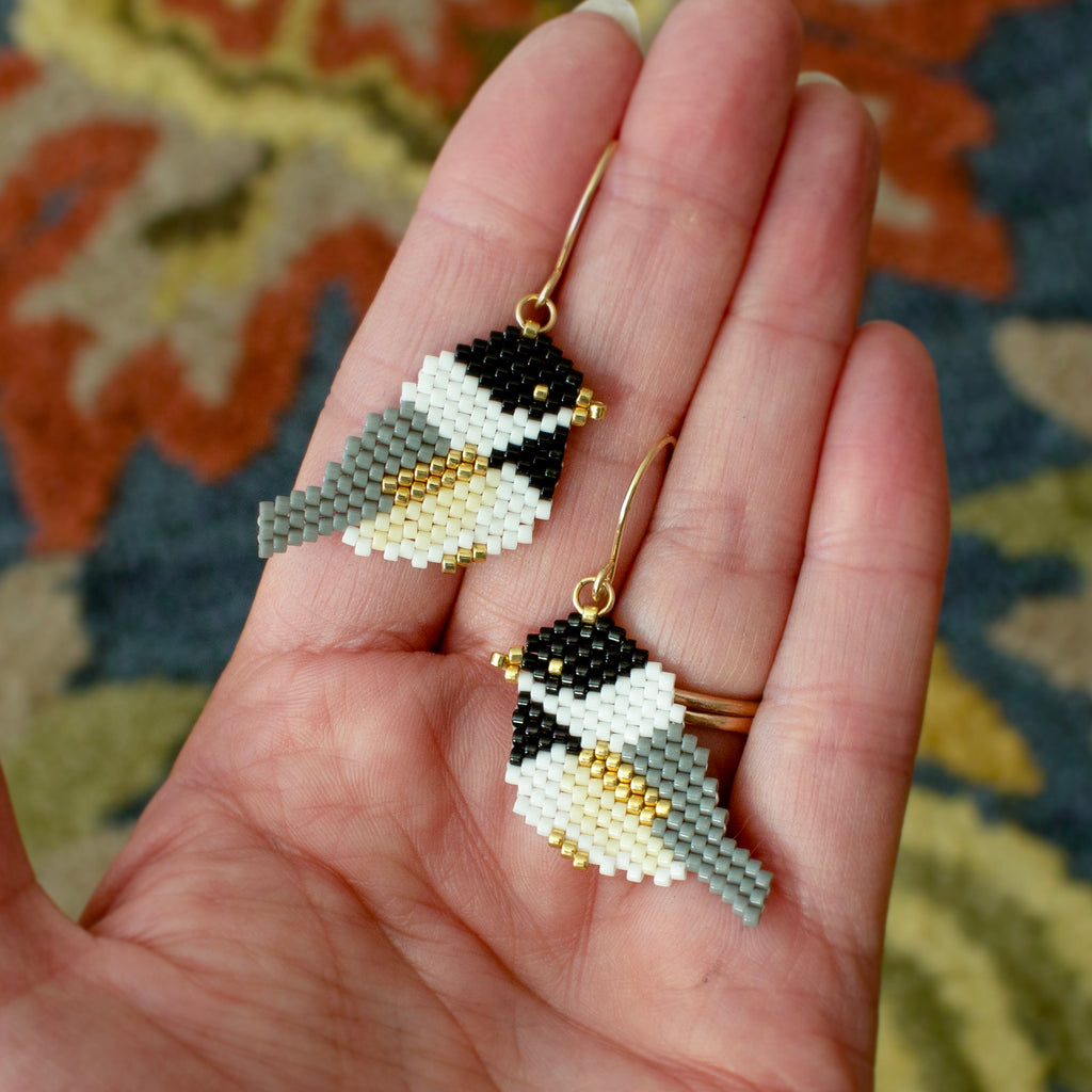 A pair of handmade beaded bird drop earrings with gold hooks, held in a person's hand. The earrings are detailed with black, white, gray, and gold seed beads, set against a patterned rug background.