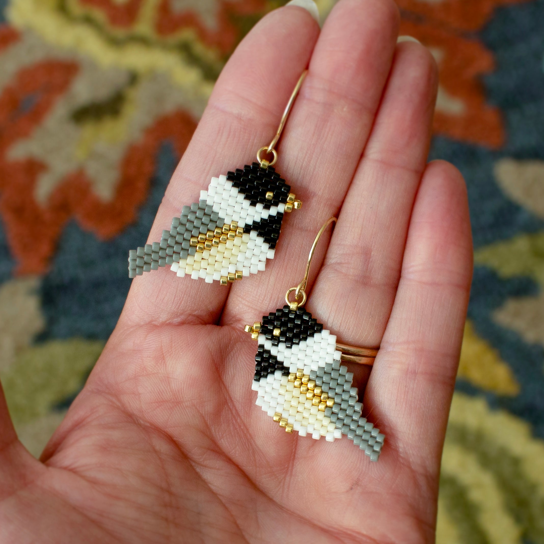 A pair of handmade beaded bird drop earrings with gold hooks, held in a person's hand. The earrings are detailed with black, white, gray, and gold seed beads, set against a patterned rug background.