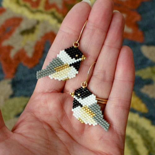 A pair of handmade beaded bird drop earrings with gold hooks, held in a person's hand. The earrings are detailed with black, white, gray, and gold seed beads, set against a patterned rug background.