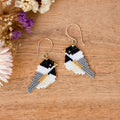 A pair of handmade beaded bird drop earrings with gold fishhook wires, displayed on a wooden surface with dried flowers in the corner.