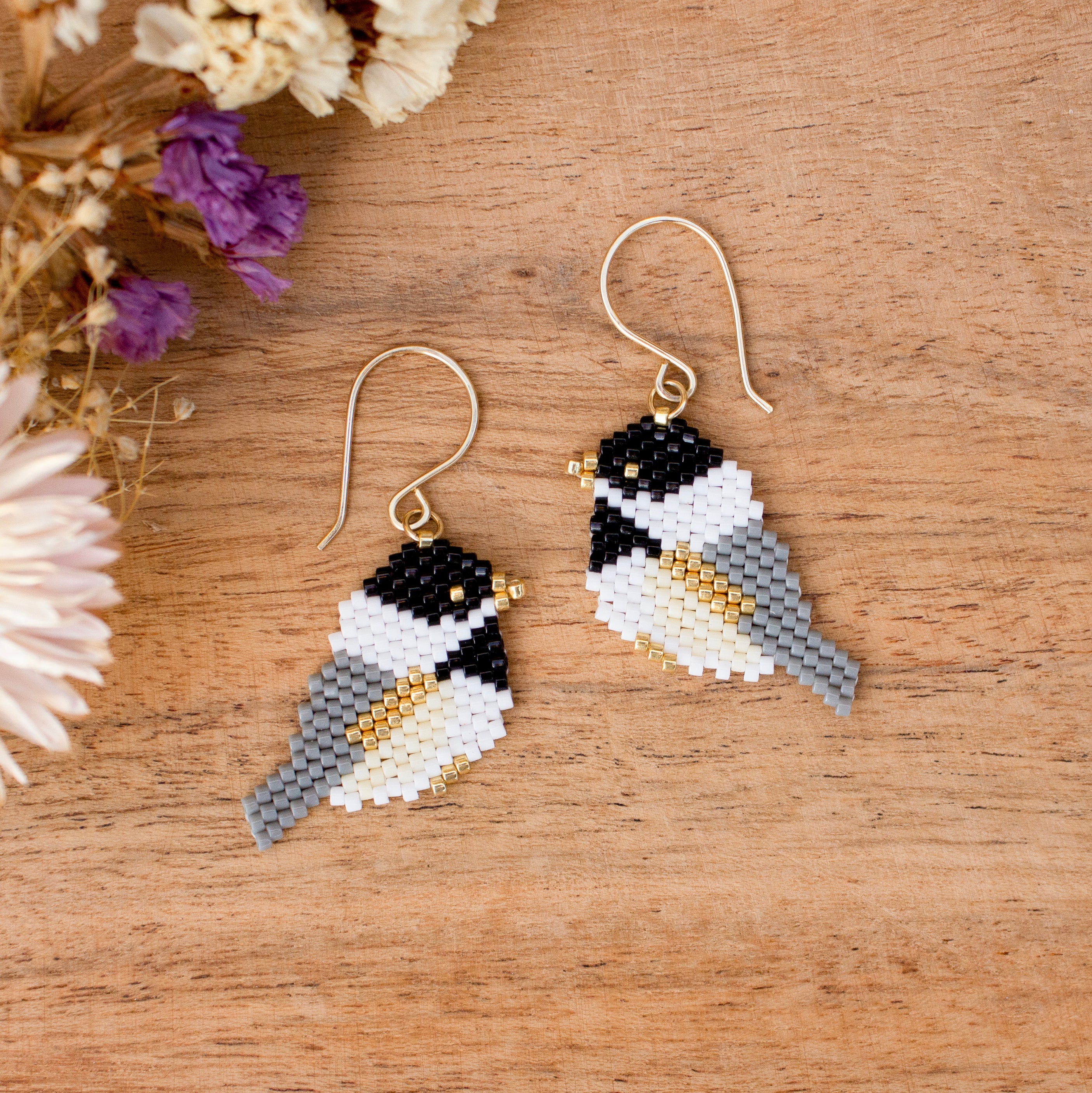 A pair of handmade beaded bird drop earrings with gold fishhook wires, displayed on a wooden surface with dried flowers in the corner.