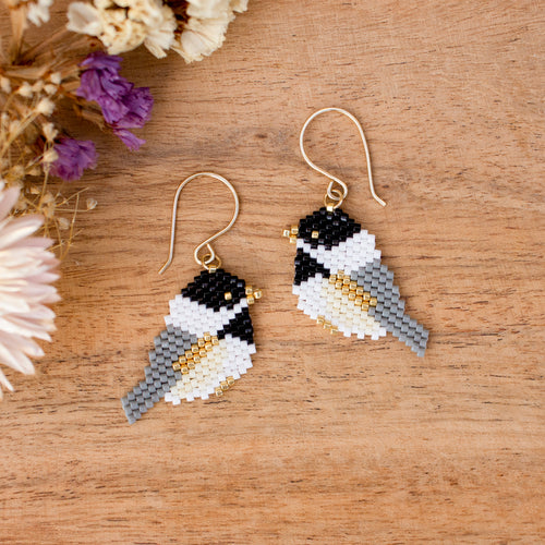A pair of handmade beaded bird drop earrings with gold fishhook wires, displayed on a wooden surface with dried flowers in the corner.