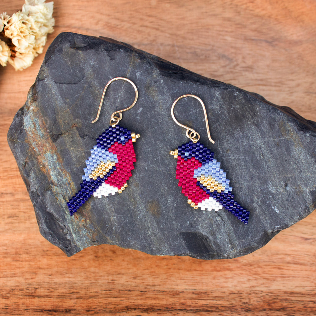 A pair of handmade beaded bird drop earrings displayed on a dark slate rock over a wooden surface. The earrings feature gold fishhook wires and beads forming birds with navy blue, red, light blue, white, and yellow details.