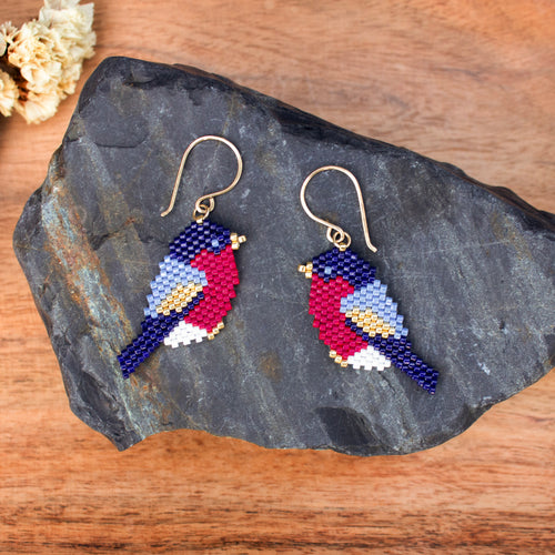 A pair of handmade beaded bird drop earrings displayed on a dark slate rock over a wooden surface. The earrings feature gold fishhook wires and beads forming birds with navy blue, red, light blue, white, and yellow details.