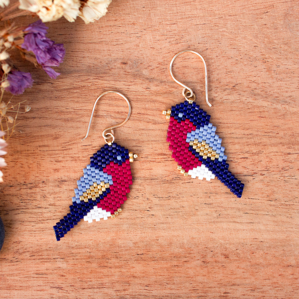 A pair of handmade beaded bird earrings on a wooden surface with dried flowers in the corner. The birds are crafted from multicolored seed beads in navy blue, red, light blue, white, and gold, hanging from gold fishhook wires.
