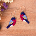 A pair of handmade beaded bird earrings on a wooden surface with dried flowers in the corner. The birds are crafted from multicolored seed beads in navy blue, red, light blue, white, and gold, hanging from gold fishhook wires.