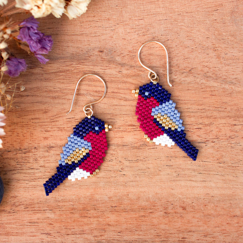 A pair of handmade beaded bird earrings on a wooden surface with dried flowers in the corner. The birds are crafted from multicolored seed beads in navy blue, red, light blue, white, and gold, hanging from gold fishhook wires.