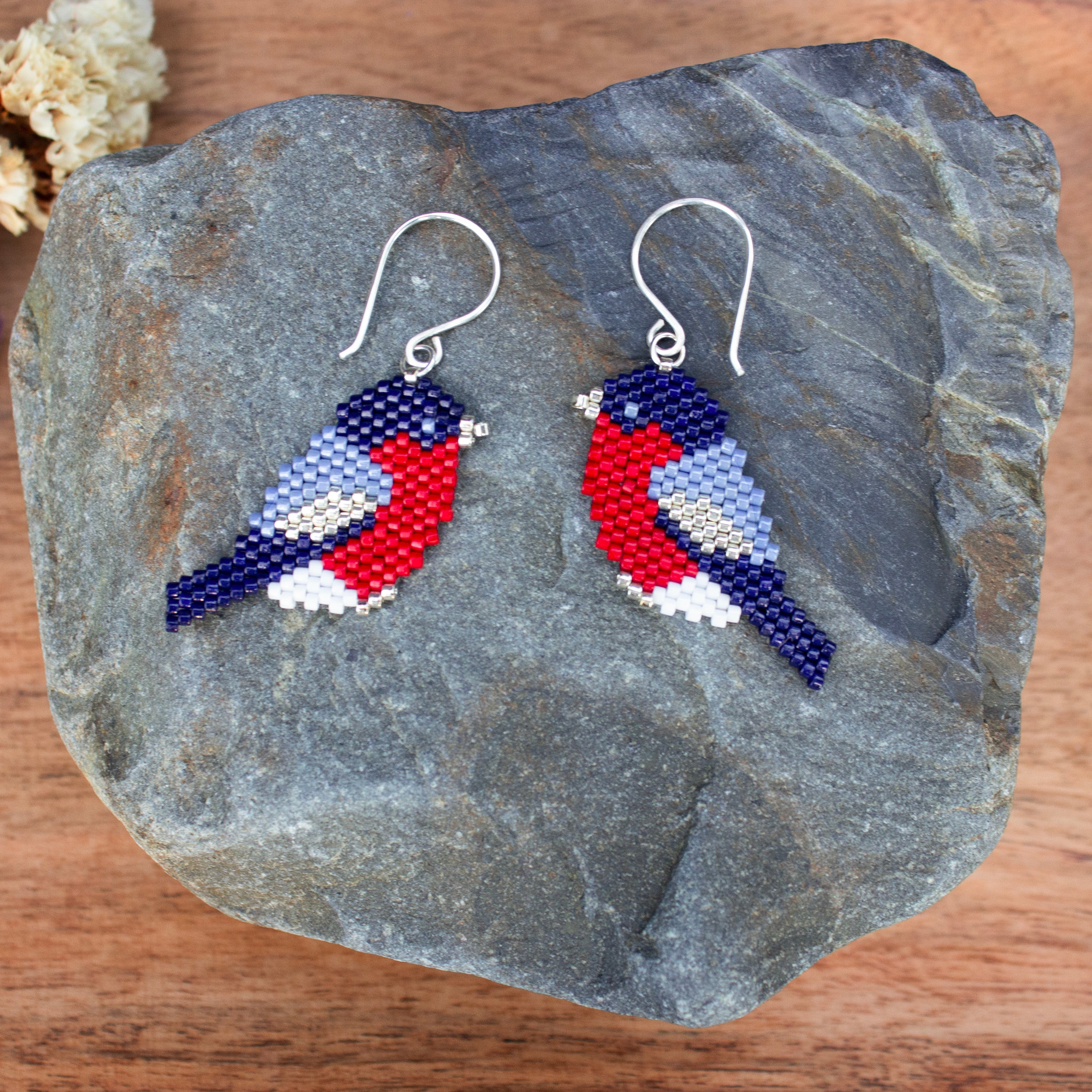 A pair of beaded bird earrings displayed on a dark gray stone over a wooden surface. The birds are made from blue, red, white, and light blue seed beads, with silver fishhook ear wires.
