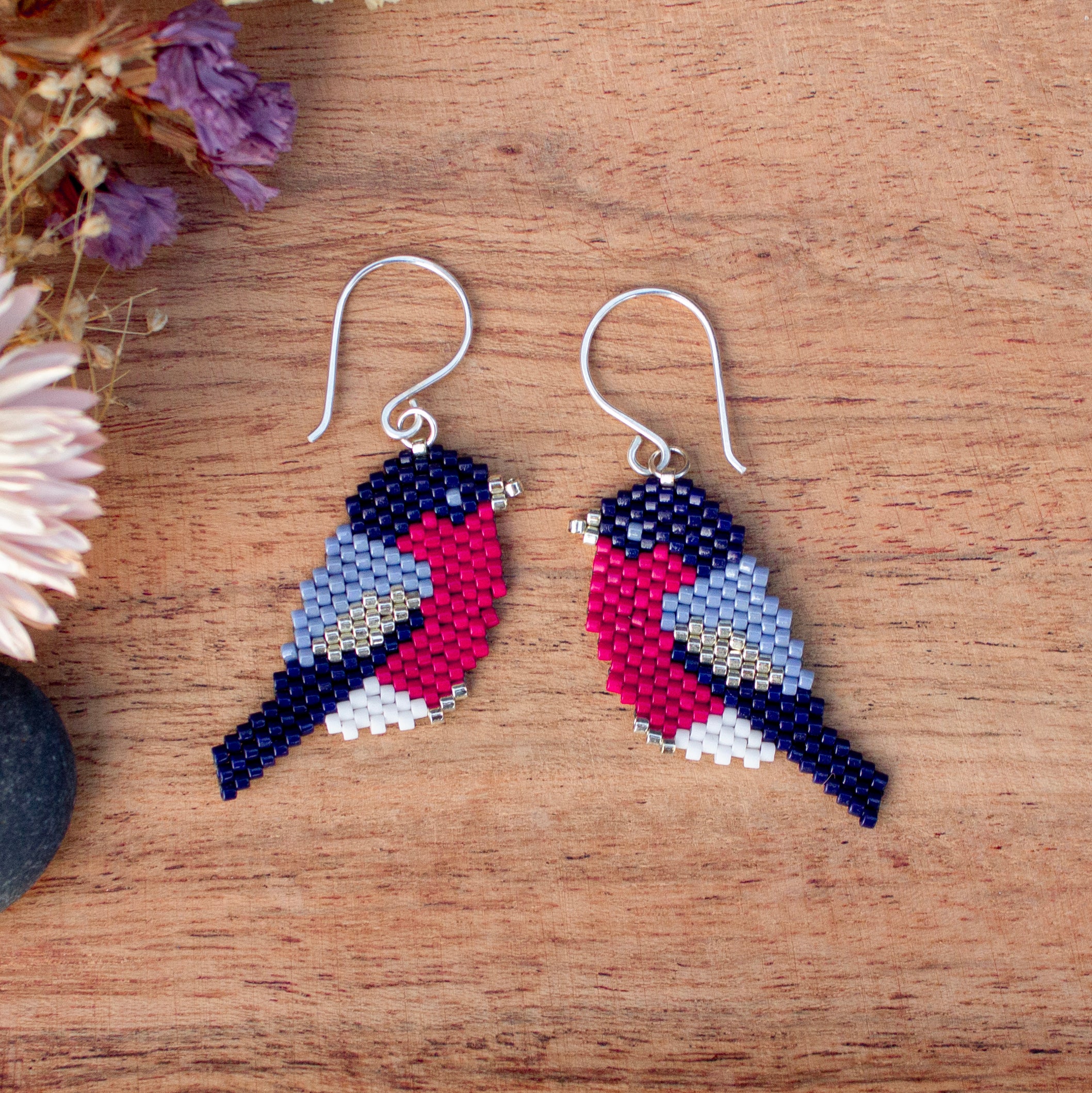 A pair of beaded bird drop earrings with silver fishhook wires, displayed on a wooden surface with dried flowers in the corner.