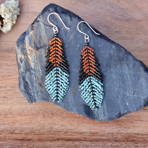 A pair of handmade beaded dangle earrings with a chevron pattern in orange, black, and light blue, displayed on a dark slate rock over a wooden surface.